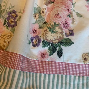 May include: Close-up of a floral fabric featuring large pink, purple, and yellow roses on a cream background. The fabric has a red and white checkered border, and a green and white striped bottom edge. The overall theme is vintage floral.