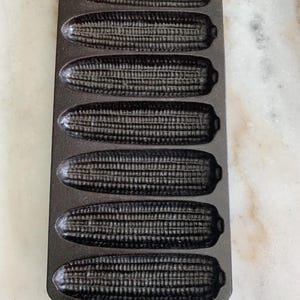 May include: A black cast iron cornbread pan shaped like seven ears of corn. The pan has a textured surface and a small hole at the top for hanging. The pan is designed for baking cornbread.