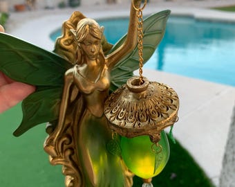 May include: A golden fairy figurine with green wings and a green dress, holding a decorative lantern. The lantern has a gold filigree top and a green glass orb with a dangling crystal. The fairy is holding the lantern with a gold chain.