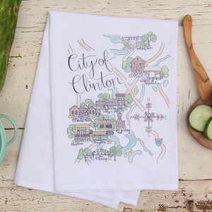 May include: A white kitchen towel with a hand-drawn map of the City of Clinton. The map features various landmarks and buildings, including a bridge, a mill, and a factory. The map is surrounded by a green border and includes a compass rose.