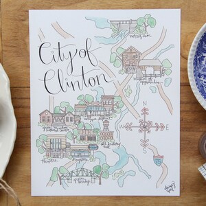 May include: A hand-drawn watercolor map of the City of Clinton, Tennessee. The map features various landmarks and points of interest, including the Green McAdoo Cultural Center, the Antique District, and the Old Jailhouse Mall. The map is surrounded by a light blue border and includes a compass rose.