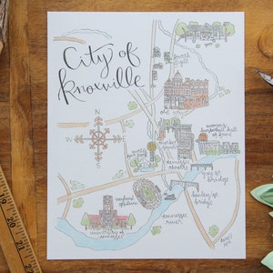 May include: A hand-drawn map of Knoxville, Tennessee, with a compass rose and labeled landmarks, including the University of Tennessee, Neyland Stadium, and the Gay Street Bridge. The map is drawn in black, brown, and blue ink on white paper.