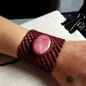 May include: A maroon crocheted bracelet with a pink stone in the center. The bracelet is on a person's wrist.