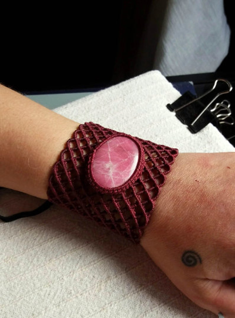May include: A maroon crocheted bracelet with a pink stone in the center. The bracelet is on a person's wrist.