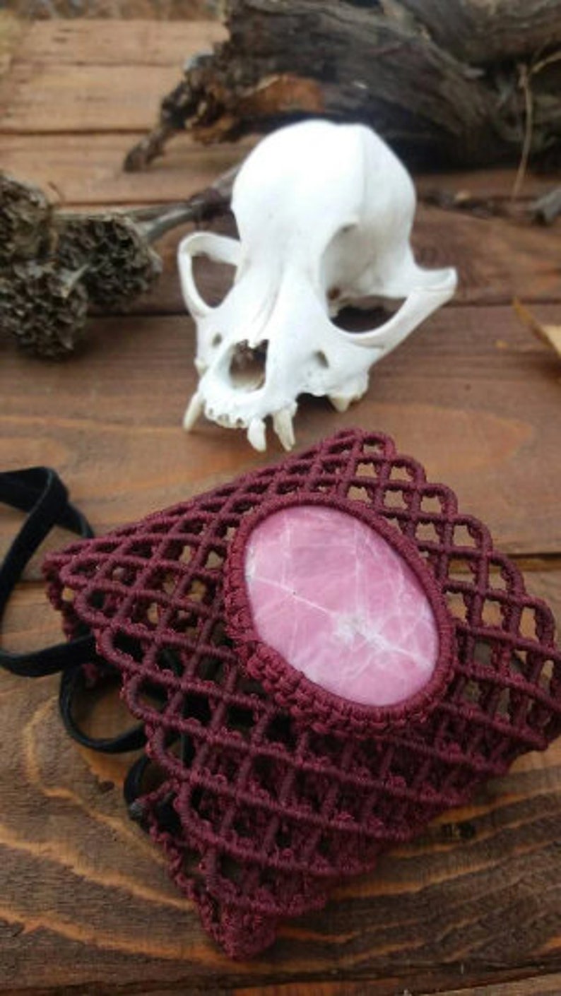 May include: A burgundy crocheted bracelet with a pink stone in the center. The bracelet is made with a grid pattern and has a delicate, feminine look.