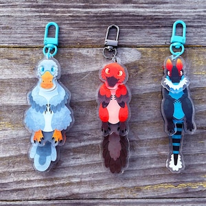 May include: Three acrylic keychains featuring cartoon-style creatures. The keychains have teal-colored clasps. One is a blue bird-like creature with an orange beak and feet. The second is a red and black creature. The third is a black and blue creature.