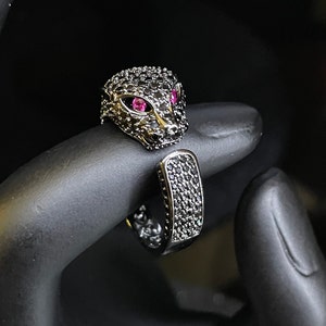 May include: A black and silver ring featuring a panther head design. The panther has pink eyes and is set with black stones.