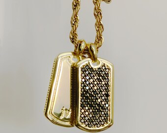Puede incluir: Un collar de cadena de oro con dos colgantes de placa de identificación de oro. Un colgante es liso y el otro está cubierto de pequeñas piedras negras.