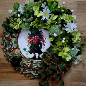 Puede incluir: Una corona verde, blanca y marrón con una ilustración de duende en el centro. La corona está decorada con flores verdes y blancas, hojas verdes y una cinta de cuadros verde y marrón.