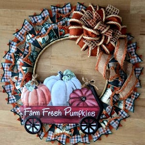 Puede incluir: Una corona de otoño con un lazo de arpillera y un carro rojo con el texto "Farm Fresh Pumpkins". El carro está decorado con tres calabazas: una naranja, una blanca y una burdeos.