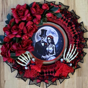 Può includere: Una ghirlanda di Halloween con uno schema di colori rosso, nero e bianco. La ghirlanda è decorata con rose rosse, pizzo nero e un'immagine rotonda di una coppia di scheletri in un bosco illuminato dalla luna. L'immagine ha il testo "Until Death Do Us Part".