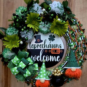 Może przedstawiać: Zielono-złoty wieniec na Święto Patryka z tabliczką z napisem "Leprechauns Welcome". Wieniec jest ozdobiony zielonymi i białymi kwiatami, zielono-złotą wstążką i dwoma gnomami z czerwonymi brodami.