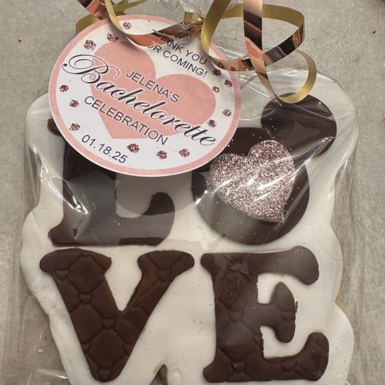 Peut inclure: Un biscuit blanc d&eacute;cor&eacute; de gla&ccedil;age au chocolat brun en forme du mot "LOVE". Un petit biscuit en forme de c&oelig;ur avec des paillettes roses est plac&eacute; sur le "O". Une &eacute;tiquette rose et blanche avec le texte "Jelena's Bachelorette Celebration 01.18.25" est attach&eacute;e au biscuit.