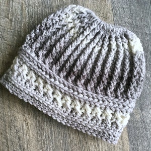 Messy bun hat displayed layed flat. Heavily textured vertical ribbing top half, wide cross stitch texture band from ear height down with horizontal ribbing at top and bottom of band.  Marled ombre in grays and off white.