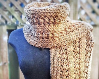 May include: A tan-colored, hand-knitted scarf featuring a textured, repeating vertical cable and rib stitch pattern. The scarf is wrapped around a dark-colored mannequin torso, displaying its length and the intricate, thick yarn construction.