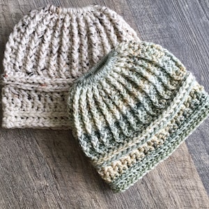 Two messy bun hat displayed layed flat, overlapping. Heavily textured vertical ribbing top half, wide cross stitch texture band with horizontal ribbing at top and bottom of band.  Marled ombre in greens (front) and cream tweed with brown fleck(back).