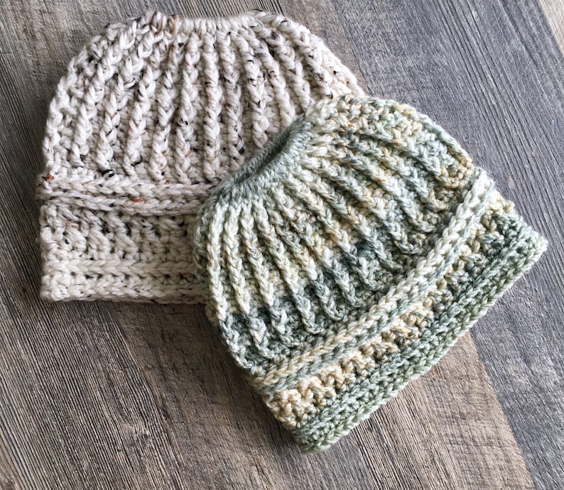 Two messy bun hat displayed layed flat, overlapping. Heavily textured vertical ribbing top half, wide cross stitch texture band with horizontal ribbing at top and bottom of band.  Marled ombre in greens (front) and cream tweed with brown fleck(back).