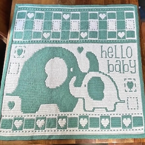 May include: A handmade baby blanket in shades of green and white. The blanket features a design of two elephants, hearts, and the words "hello baby". The blanket is square and appears to be crocheted.