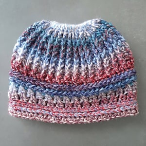 Messy bun hat displayed layed flat. Heavily textured vertical ribbing top half, wide cross stitch texture band from ear height down with horizontal ribbing at top and bottom of band.  Marled ombre in blues red and white  off white.