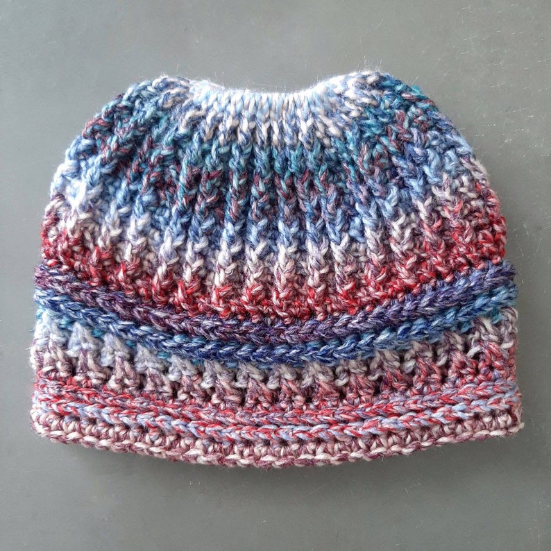 Messy bun hat displayed layed flat. Heavily textured vertical ribbing top half, wide cross stitch texture band from ear height down with horizontal ribbing at top and bottom of band.  Marled ombre in blues red and white  off white.