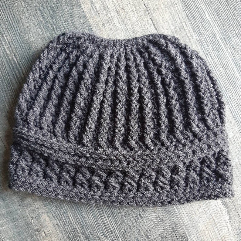 Messy bun hat displayed layed flat. Heavily textured vertical ribbing top half, wide cross stitch texture band from ear height down with horizontal ribbing at top and bottom of band.  Solid slate gray.
