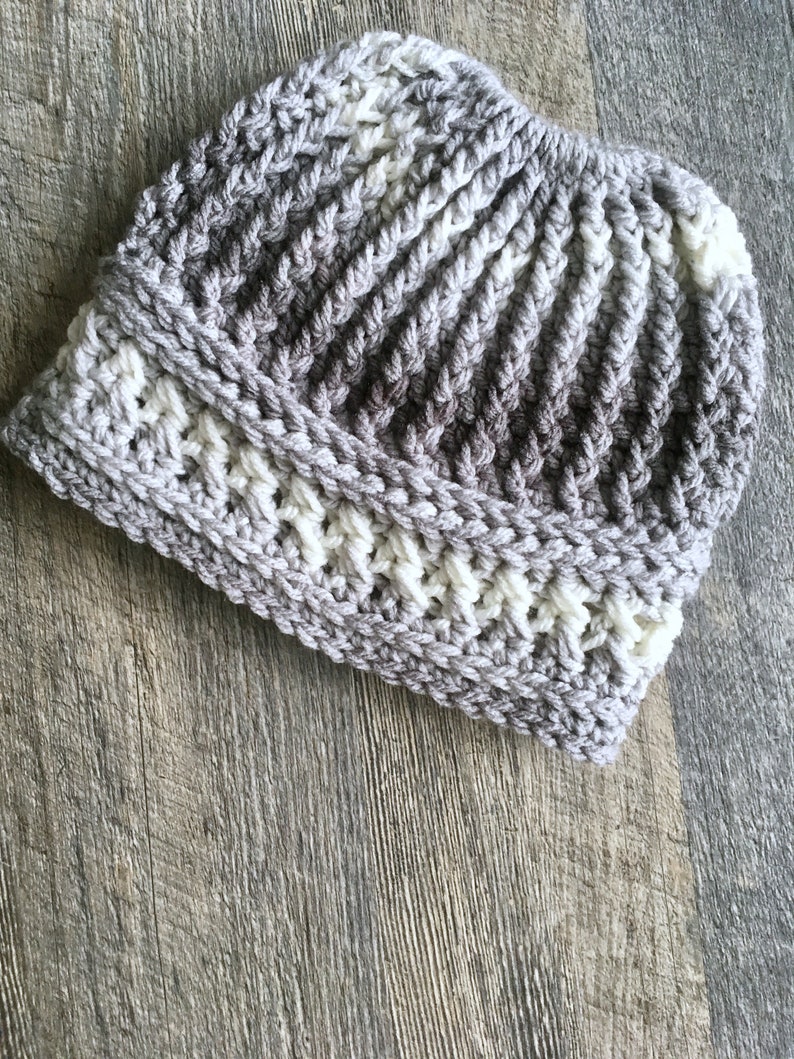 Messy bun hat displayed layed flat. Heavily textured vertical ribbing top half, wide cross stitch texture band from ear height down with horizontal ribbing at top and bottom of band.  Marled ombre in grays and off white.