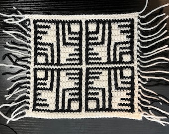 May include: A black and white crocheted square with a geometric pattern and fringe on all sides.