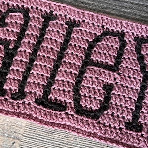 Op de afbeelding: Close-up van een gehaakt item in een stoffige roze kleur. Het woord "SALLY" is met zwart garen op het item gehaakt. De steken zijn zichtbaar en creëren een textuur.