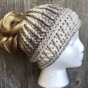 Messy bun hat with top pony tail displayed on a mannequin head. Heavily textured vertical ribbing top half, wide cross stitch texture band from ear height down with horizontal ribbing at top and bottom of band.  Marled ombre in brown, tan, off white.