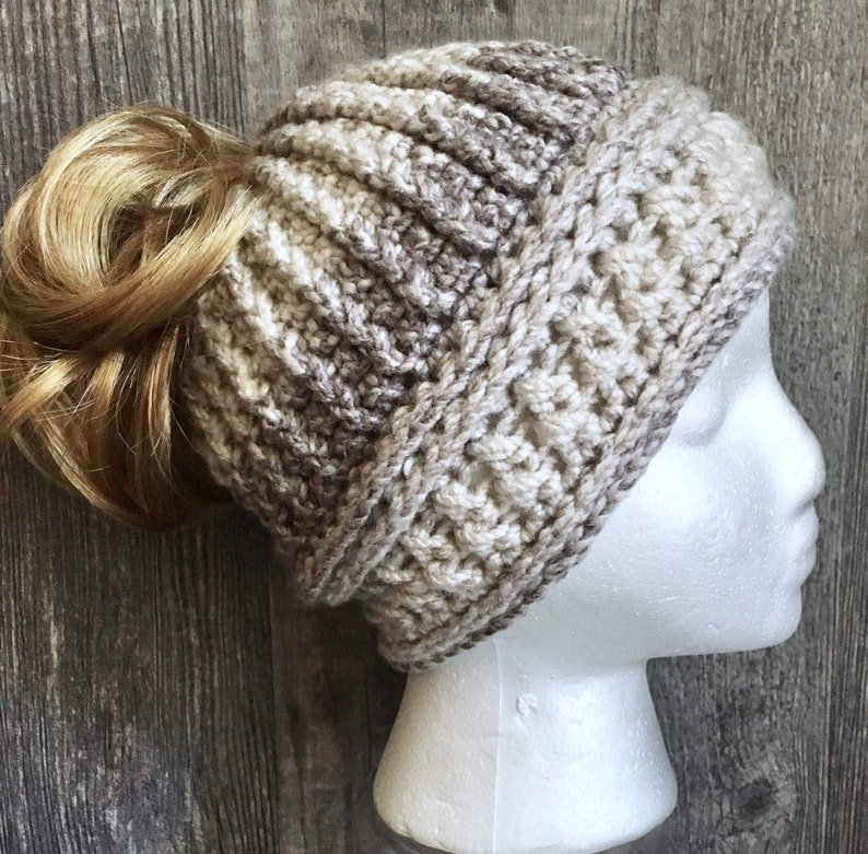 Messy bun hat with top pony tail displayed on a mannequin head. Heavily textured vertical ribbing top half, wide cross stitch texture band from ear height down with horizontal ribbing at top and bottom of band.  Marled ombre in brown, tan, off white.
