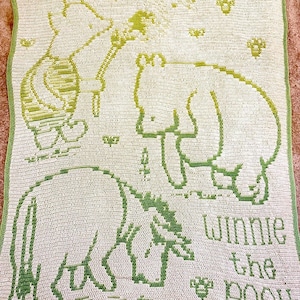Puede incluir: Una manta de ganchillo rectangular en color crema y verde claro con un diseño gráfico de los personajes de Winnie the Pooh. El patrón incluye a Piglet, Winnie the Pooh y Eeyore, rodeados de pequeñas abejas, con el texto Winnie the Pooh.