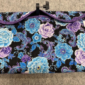 May include: A quilted, padded sewing machine cover with a black background and a floral pattern in shades of blue and purple. The cover has a purple border and a black adjustable handle.