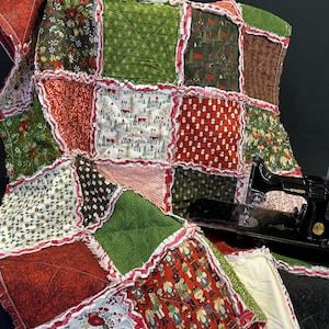 Puede incluir: Un edredón navideño hecho a mano con un diseño de patchwork. El edredón presenta varias telas en rojo, verde y blanco, con patrones festivos como flores de Nochebuena, casas cubiertas de nieve y cascanueces. Los bordes están adornados con un borde rojo con volantes.