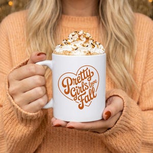 May include: A white ceramic mug featuring a heart-shaped graphic in burnt orange with the text Pretty Girls love Fall. The mug is filled with a beverage topped with a generous swirl of whipped cream and a light dusting of brown spice sprinkles.