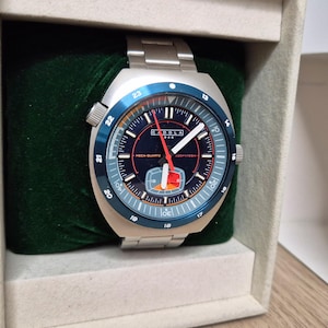 Könnte beinhalten: Eine silberfarbene Armbanduhr mit blauer Lünette und schwarzem Zifferblatt. Die Uhr hat weiße und rote Zeiger, und der Text "CADOLA" ist sichtbar. Die Uhr wird in einer grün mit Samt ausgekleideten Box präsentiert.