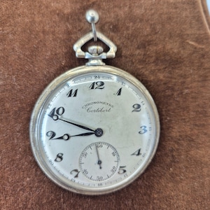 May include: Silver pocket watch with a white face, featuring black numerals and hands. The watch has a subdial at the bottom and the text "CHRONOMETRE Cortébert" is visible. The watch is a classic timepiece.