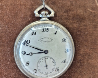 Peut inclure: Montre de poche argentée avec un cadran blanc, des chiffres et des aiguilles noirs. La montre a un sous-cadran en bas et le texte "CHRONOMETRE Cortébert" est visible. Une montre classique.