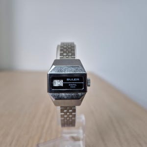 Könnte beinhalten: Eine silberfarbene digitale Buler-Uhr mit rechteckigem Zifferblatt und Gliederarmband. Die Uhr zeigt die Uhrzeit 1:35 an. Das Wort "Buler" ist über der Digitalanzeige und "Digital Swiss" darunter aufgedruckt.