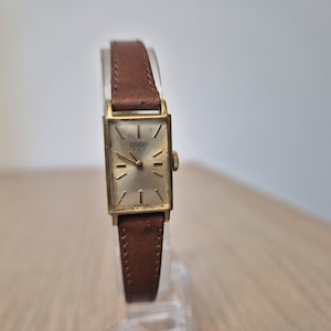 Op de afbeelding: Een vintage rechthoekig polshorloge met een goudkleurige kast en een lichtgekleurde wijzerplaat. Het horloge heeft een bruine leren band. De wijzerplaat toont de merknaam "Gevril". Het horloge wordt op een transparante standaard getoond.