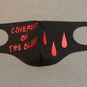 May include: Black face mask with the words "COVERED BY THE BLOOD" in red lettering. The mask also features three red teardrop shapes on the right side. The mask is made of a soft, flexible material.