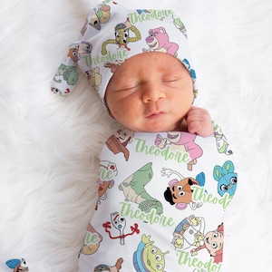 May include: A white swaddle blanket with a repeating pattern of Toy Story characters, including Woody, Buzz Lightyear, and Jessie. The blanket is personalized with the name "Theodore" in green text. A matching hat with the same pattern and personalization is also shown.