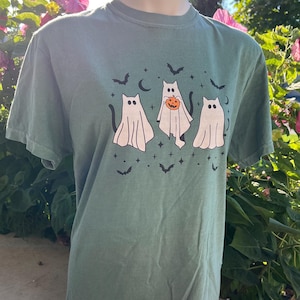 May include: A green t-shirt with a Halloween design. The graphic shows three ghost-cat figures, bats, stars, and a crescent moon. One ghost-cat holds a pumpkin. The shirt is displayed on a mannequin.