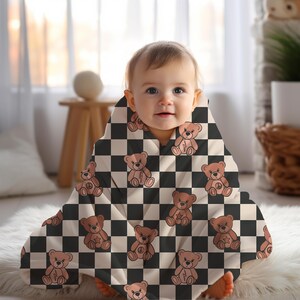 Pu&ograve; includere: Un bambino che indossa una copertina per neonati a scacchi bianchi e neri con un motivo di orsacchiotto di peluche marrone. La copertina ha un orsacchiotto di peluche marrone con un fulmine sul petto su ogni quadrato.