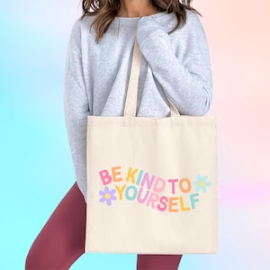 May include: Cream-colored tote bag with the text "BE KIND TO YOURSELF" in colorful, playful lettering. The bag features a floral design and is held by a person wearing a gray sweater and maroon leggings.