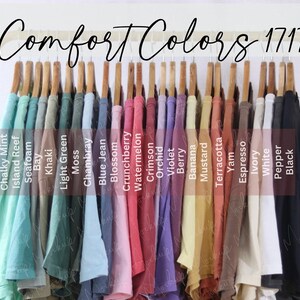 May include: A selection of Comfort Colors 1717 t-shirts in a range of hues, such as Chalky Mint, Island Reef, and Black. The shirts are displayed on wooden hangers on a gold rack. The text "Comfort Colors 1717" is visible.