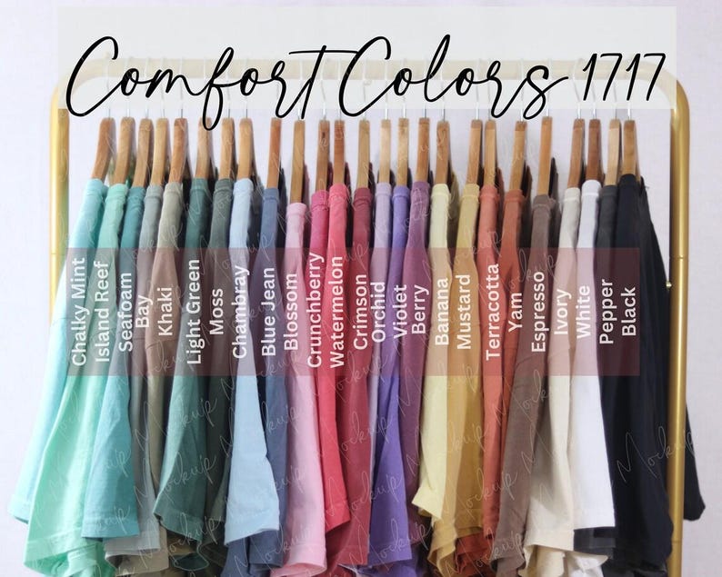 May include: A selection of Comfort Colors 1717 t-shirts in a range of hues, such as Chalky Mint, Island Reef, and Black. The shirts are displayed on wooden hangers on a gold rack. The text "Comfort Colors 1717" is visible.