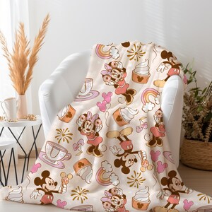 May include: A soft, cream-colored throw blanket with a repeating pattern of Mickey Mouse and Minnie Mouse characters. The characters are depicted in a vintage cartoon style with pink, red, and yellow accents. The blanket features a whimsical design with ice cream cones, teacups, and fireworks.