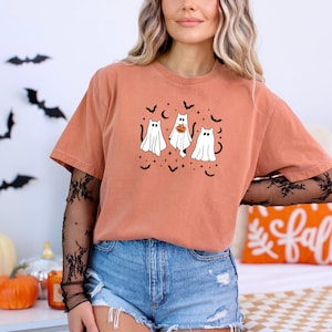 May include: An orange t-shirt with a Halloween design. The graphic shows three ghost-cat figures, bats, and stars. The shirt is worn with denim shorts. The sleeves are short.