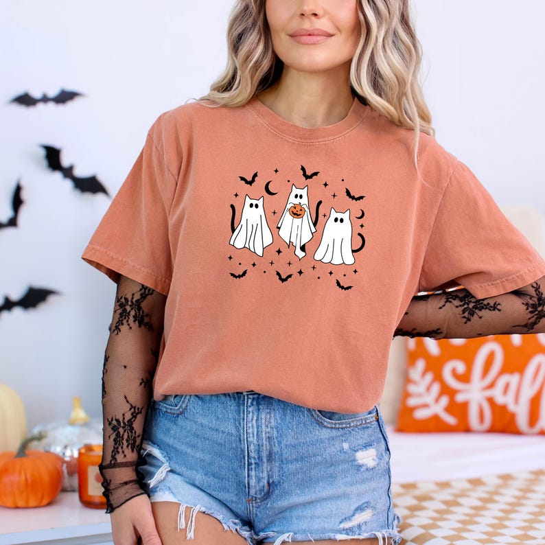 May include: An orange t-shirt with a Halloween design. The graphic shows three ghost-cat figures, bats, and stars. The shirt is worn with denim shorts. The sleeves are short.