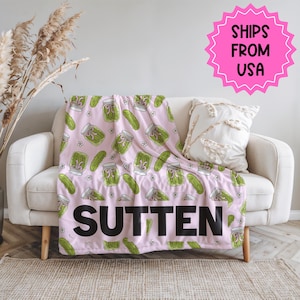 May include: A pink blanket featuring a pattern of green pickle jars and pickles, with small white flowers. The word "SUTTEN" is printed in large black letters. The blanket is draped over a white sofa.